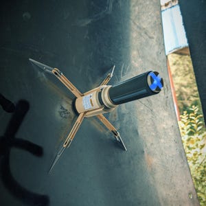 May include: A beige and black tool with four silver prongs extending outward, attached to a dark surface. The tool has a textured black grip and a blue cross design on the end. The tool appears to be a specialized device.