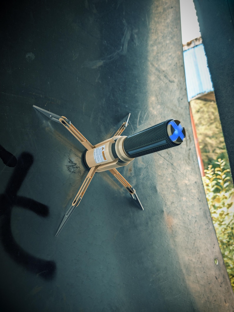 May include: A beige and black tool with four silver prongs extending outward, attached to a dark surface. The tool has a textured black grip and a blue cross design on the end. The tool appears to be a specialized device.