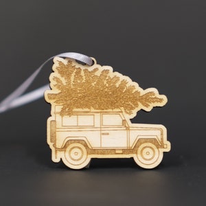 May include: A wooden ornament depicting a vintage car with a Christmas tree on top. The ornament is painted in a light brown colour and has a white string attached to it.