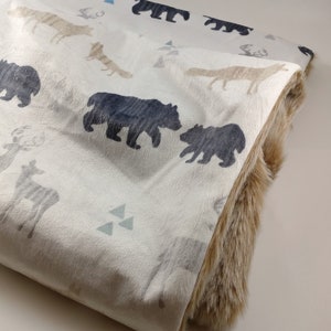 May include: A white and beige fleece blanket with a brown faux fur trim. The blanket features a repeating pattern of black bears, deer, foxes, and triangles.