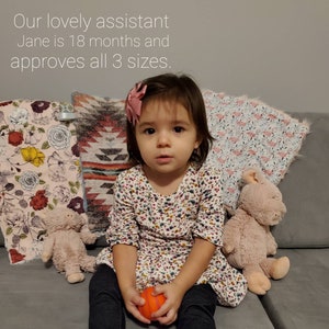 K&ouml;nnte beinhalten: Ein junges M&auml;dchen mit einer rosa Schleife im Haar sitzt auf einem Sofa mit einem Pl&uuml;schtier. Sie tr&auml;gt ein wei&szlig;es Kleid mit Blumenmuster und eine schwarze Hose. Sie h&auml;lt einen orangenen Ball in ihren H&auml;nden. Das Sofa ist mit einem grauen Stoff bezogen. Hinter ihr befinden sich drei Kissen, eines mit einem Blumenmuster, eines mit einem geometrischen Muster und eines mit einem rosa und wei&szlig;en Muster.