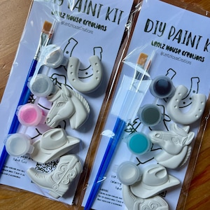 May include: Two DIY paint kits in clear plastic packaging, each containing a blue paintbrush, three small paint pots, and three white ceramic figures: a horseshoe, a horse head, and a cowboy boot. The backing card reads DIY PAINT KIT.