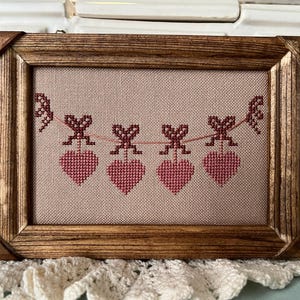May include: A framed cross-stitch design featuring a string of hearts with bows hanging from it. The hearts are red and the bows are brown. The background is a light beige.