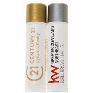 May include: Two white tubes of lip balm with gold and silver lids. The left tube has a gold logo with the text "(C) 21 CENTURY 21" and "Dynamic Realty" below. The right tube has a red and black logo with the text "KW GREATER CLEVELAND NORTHEAST" and "KELLERWILLIAMS." below.