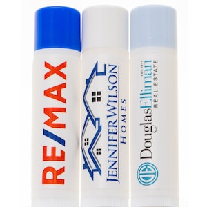 May include: Three white tubes of lip balm with different real estate company logos. The first tube has a blue cap and the logo "RE/MAX" in red and white. The second tube has a white cap and the logo "Jennifer Wilson Homes" in blue and white. The third tube has a light blue cap and the logo "Douglas Elliman Real Estate" in black and white.