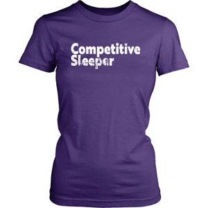 May include: A purple t-shirt with the words "Competitive Sleeper" printed in white, distressed font. The shirt has short sleeves and a crew neck.
