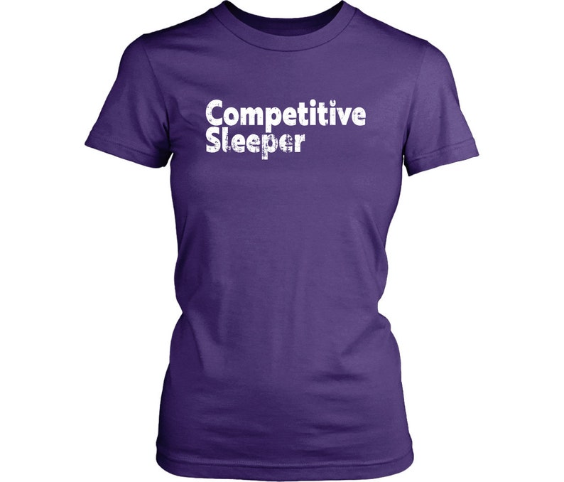 May include: A purple t-shirt with the words "Competitive Sleeper" printed in white, distressed font. The shirt has short sleeves and a crew neck.