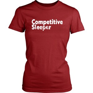 May include: A red t-shirt with the words "Competitive Sleeper" printed in white. The text has a distressed, textured appearance. The shirt has short sleeves and a crew neck.