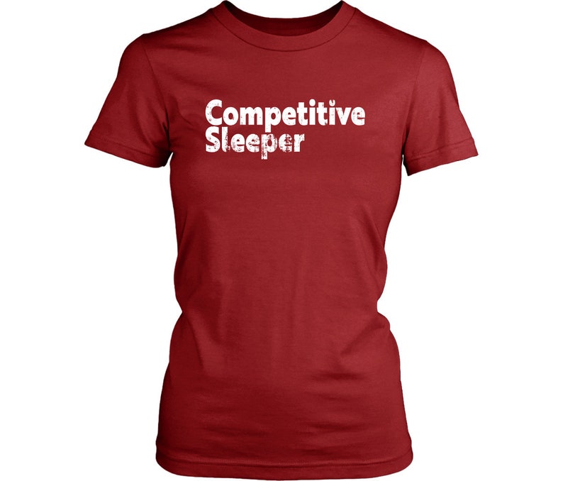 May include: A red t-shirt with the words "Competitive Sleeper" printed in white. The text has a distressed, textured appearance. The shirt has short sleeves and a crew neck.