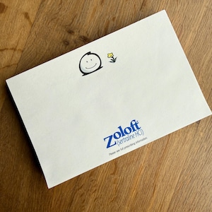 May include: A white card with a smiling face and a small yellow flower illustration. The word "Zoloft" is printed in blue, along with the chemical name and a disclaimer. The card is on a wooden surface.