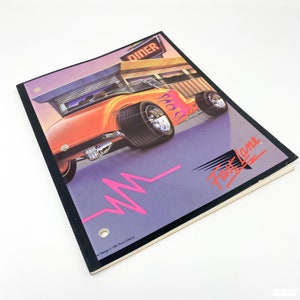 May include: A vintage-style notebook with a black cover and a colorful illustration of a hot rod car driving past a diner. The notebook has a pink and white heart beat line and the text "Fast Lane" in a stylized font.