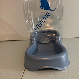 May include: A clear plastic pet food and water dispenser with a blue silhouette of a dog and the word "STERLING" printed on the side. The dispenser has a gray plastic base with a bowl for water or food.