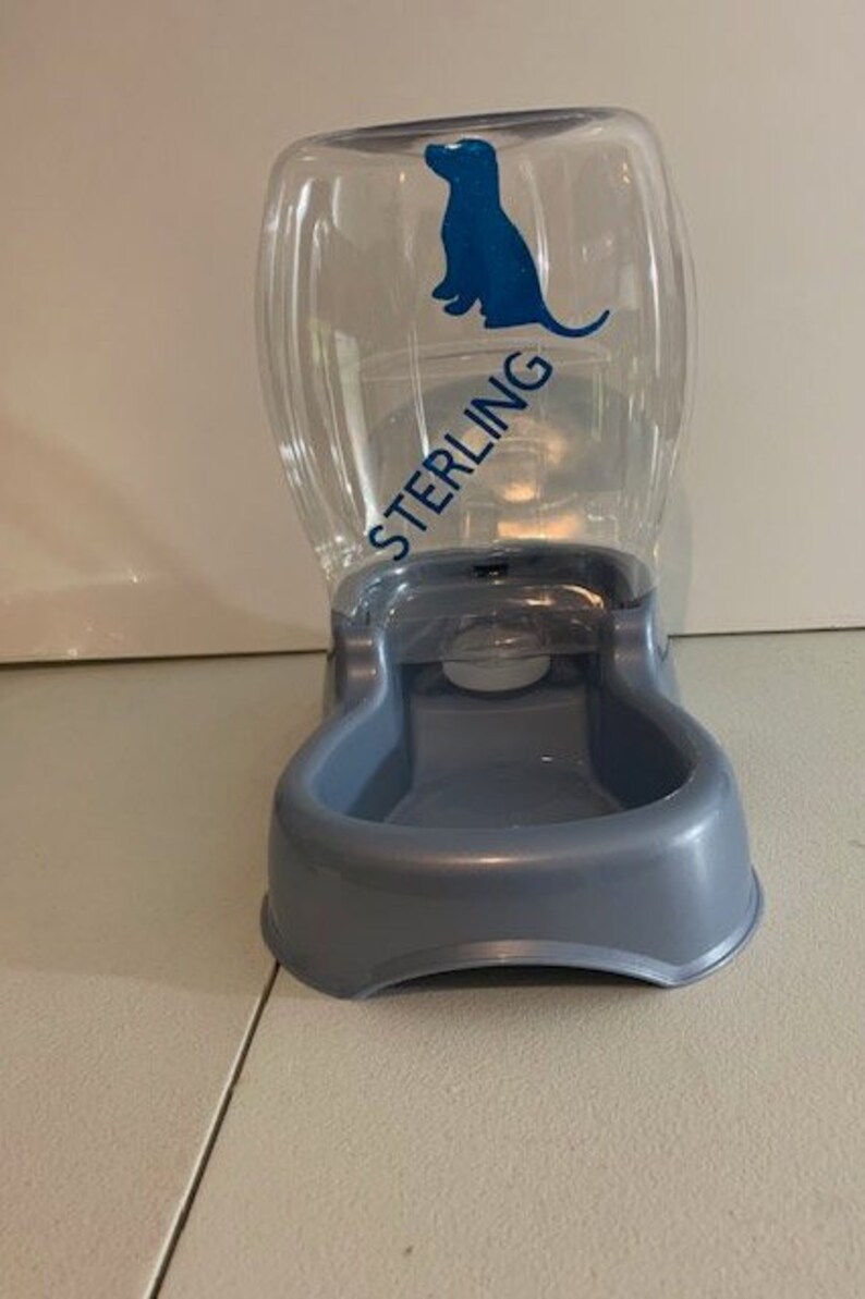 May include: A clear plastic pet food and water dispenser with a blue silhouette of a dog and the word "STERLING" printed on the side. The dispenser has a gray plastic base with a bowl for water or food.