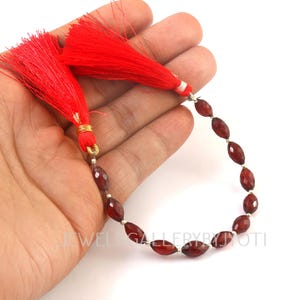 May include: A strand of faceted, oval-shaped, dark red beads with silver accents, connected to two bright red tassels. The beads are held in a hand against a white background. The text "JEWEL GALLERY BY JYOTI" is visible.