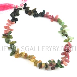 May include: A strand of multi-colored tourmaline gemstone beads. The beads are in a graduated color pattern, starting with black, then green, pink, and yellow. The beads are all rough and have a natural, unpolished look. The strand is finished with a pink tassel.