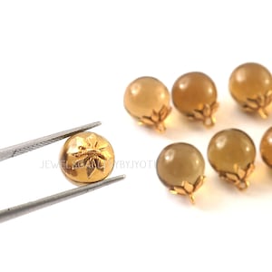 May include: Close-up of several gold-colored, round beads with a floral design. One bead is held by tweezers. The beads have a small, gold-colored base. The text "JEWELSCAN BYJYOTI" is visible.