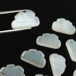 Puede incluir: Piedras preciosas en forma de nube, de color azul pálido, sobre un fondo negro. Una es sujetada por pinzas, otras están dispersas. El texto "JEWELSGALLERYBYJYOTI" es visible. Adecuado para joyería.