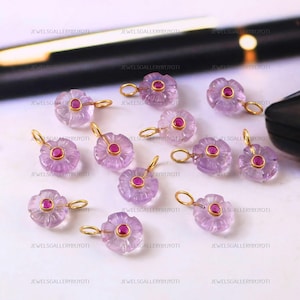 May include: A collection of amethyst flower pendants with gold-colored loops and a small red gemstone center. The pendants are carved with petal details and are displayed on a light-colored surface.