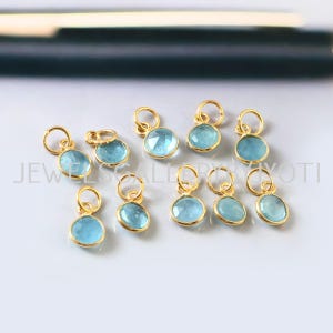 May include: Ten gold-plated charms with light blue gemstone accents. Each charm features a round gemstone with a smooth, polished finish. The charms are perfect for adding a touch of elegance to jewelry projects.