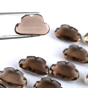 May include: A group of seven smoky quartz gemstones carved into cloud shapes. The gemstones are translucent and have a light brown color. The gemstones are arranged in a row on a white surface.