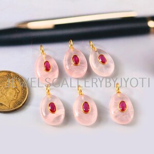 May include: Six teardrop-shaped pendants with a pinkish hue, each featuring a central oval-shaped ruby stone set in a gold-colored bezel. The pendants have a small gold loop at the top. A gold coin is visible on the left side of the image.