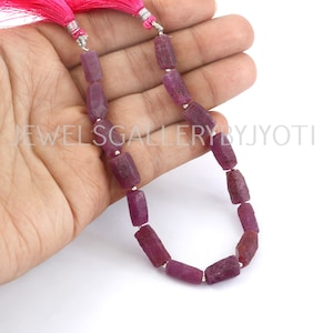 Könnte beinhalten: Eine Kette aus tiefrosa Rubinperlen, jede Perle ist facettiert und rechteckig geformt. Die Perlen sind auf einem Silberfaden aufgefädelt und haben einen rosa Quasten an der Spitze.