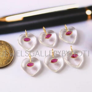 May include: Six heart-shaped pendants with a clear, glass-like appearance. Each pendant features a small, oval-shaped, ruby-colored stone and a gold-colored loop for hanging. The pendants are arranged on a light-colored surface.