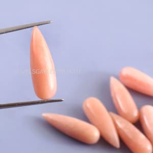 Puede incluir: Un grupo de piedras preciosas rosadas en forma de lágrima, algunas se sostienen con pinzas. JEWELSGALLERYBYJYOTI está impreso en el fondo.