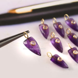 Può includere: Primo piano di diversi ciondoli in ametista con anelli color oro e piccole gemme trasparenti. I ciondoli sono di colore viola intenso e hanno la forma di punte di freccia. Un ciondolo è tenuto da una pinzetta. Il testo "JEWELSGALLERYBYJYOTI" è visibile.