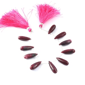 May include: A strand of 10 teardrop-shaped gemstone beads in a deep pink and red color with silver wire connecting them. The beads are strung on a silver wire with pink tassels on each end.