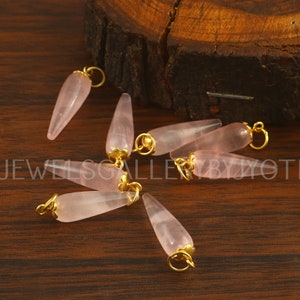 May include: Eight pink quartz teardrop pendants with gold-toned caps and jump rings. The pendants are scattered on a wooden surface.