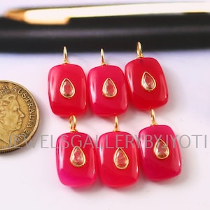 May include: Six rectangular, fuchsia-coloured gemstone pendants with gold-coloured bails. Each pendant features a teardrop-shaped, light-coloured stone in the centre. The pendants are displayed on a white surface with a gold coin in the background.