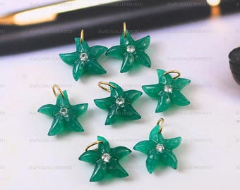 May include: Seven green star-shaped pendants with gold loops and a small, clear gem in the center of each. The pendants are arranged on a white surface, with a pen in the background.