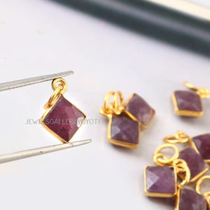 May include: Gold-plated square pendants with faceted, deep red gemstones. The pendants are held by tweezers, with several others scattered nearby. The text "JEWELS GALLERY BY JYOTI" is visible in the image.