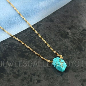 Puede incluir: Un delicado collar de cadena dorada con un pequeño colgante de piedra turquesa irregular. El collar se muestra sobre una superficie oscura y texturizada. El texto "JEWELSGALLERYJYOTI" es visible en el fondo.