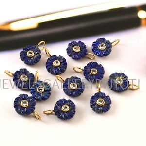 Op de afbeelding: Een verzameling donkerblauwe bloemvormige hangers met goudkleurige lussen. Elke hanger heeft een kleine, heldere steen in het midden. De hangers zijn gerangschikt op een wit oppervlak.