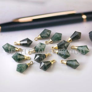 Puede incluir: Una colección de colgantes facetados en forma de lágrima de piedra preciosa verde, con tapas y bucles de metal dorado. Los colgantes están dispersos sobre una superficie blanca, con un bolígrafo en el fondo. Para la creación de joyas.