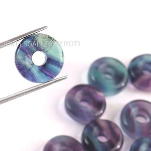Può includere: Primo piano di una perla a ciambella di fluorite blu, verde e viola con un foro al centro. La perla è tenuta da una pinzetta. Ci sono diverse altre perle a ciambella di fluorite sullo sfondo.
