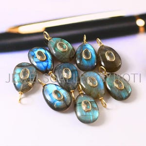 Pode incluir: Uma coleção de pingentes ovais de labradorita com argolas douradas. Os pingentes exibem uma mistura de tons azuis, verdes e cinzentos, com uma pedra menor em forma oval no centro. Os pingentes estão dispostos sobre uma superfície branca.