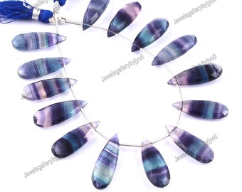 May include: A strand of 15 teardrop-shaped fluorite gemstone beads. The beads are a mix of purple, blue, and green colors with white streaks. The beads are strung on a silver wire.