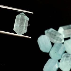 May include: A close-up of a light blue aquamarine gemstone, faceted and cut into a rectangular shape. The gemstone is held by tweezers and surrounded by other aquamarine gemstones.