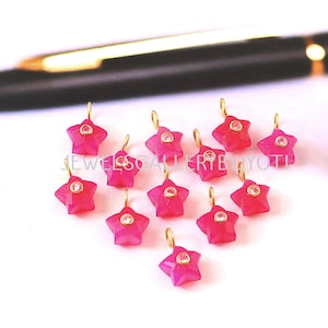 May include: A cluster of pink star-shaped charms, each with a gold-coloured loop and a small, clear gem at the centre. The charms are displayed on a white surface, with a pen visible in the background.