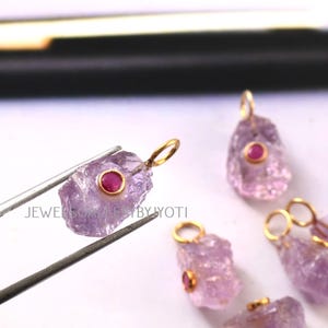 May include: Close-up of several rough-cut amethyst pendants with gold-colored bails and small, round, red gemstones. One pendant is held by tweezers. The text "JEWELSCALERYBYYOTI" is visible.