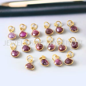 May include: A selection of gold pendants, each with a teardrop-shaped, deep pink gemstone. The gemstones are polished and set in a gold bezel. The pendants are arranged on a light surface. The gemstones are a vibrant shade of pink.