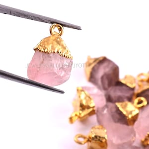 Könnte beinhalten: Eine Gruppe von rosa Rosenquarz-Anhängern mit vergoldeten Metallkappen und Sprungringen. Die Anhänger sind unregelmäßig geformt und haben ein natürliches, rohes Aussehen.