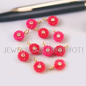 Pode incluir: Uma coleção de pequenos pingentes vermelhos em forma de flor com laços dourados. Cada pingente apresenta uma pedra preciosa transparente no centro. Os pingentes estão espalhados em uma superfície branca. O texto "JEWEL SGALLERY JYOTI" é visível.