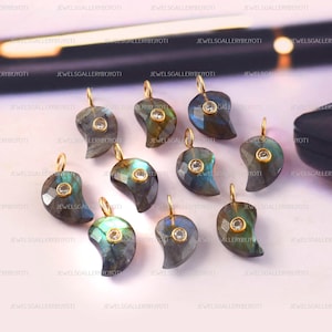 Puede incluir: Una colección de colgantes de labradorita en forma de lágrima con aros dorados y pequeñas gemas transparentes. Los colgantes tienen una mezcla de tonos azules, verdes y grises. Las joyas se exhiben sobre una superficie clara.