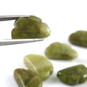 Pode incluir: Uma coleção de seis cabochões de pedras preciosas verdes, alguns dos quais têm a forma de folhas. Os cabochões estão dispostos sobre uma superfície branca. Os cabochões são lisos e polidos.
