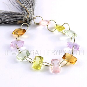 Può includere: Una collana di perle di pietre preziose sfaccettate in tonalità rosa, giallo, verde e viola. Le perle sono infilate su una catena dorata con piccoli anelli di giunzione. La catena ha un fiocco ad un'estremità.