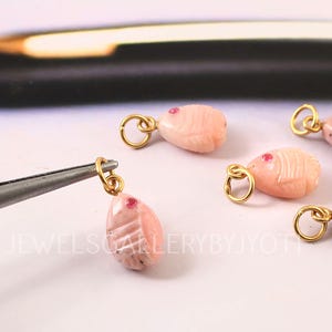 May include: Close-up of several pink gemstone charms with gold-coloured loops. Each charm is carved with a fish-like design and features a small red accent. One charm is held by a pair of tweezers.
